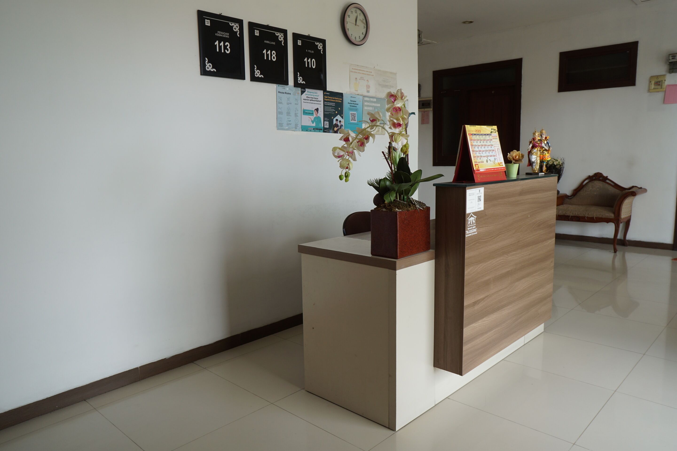 A reception desk with a clock, a potted orchid, and a small statue. There are framed notices on the wall and a wooden chair in the background.