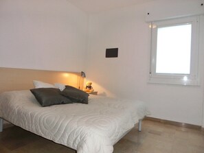 Une chambre à coucher avec un lit, des oreillers, une table de chevet avec une lampe, une fenêtre et un interrupteur fixé au mur.