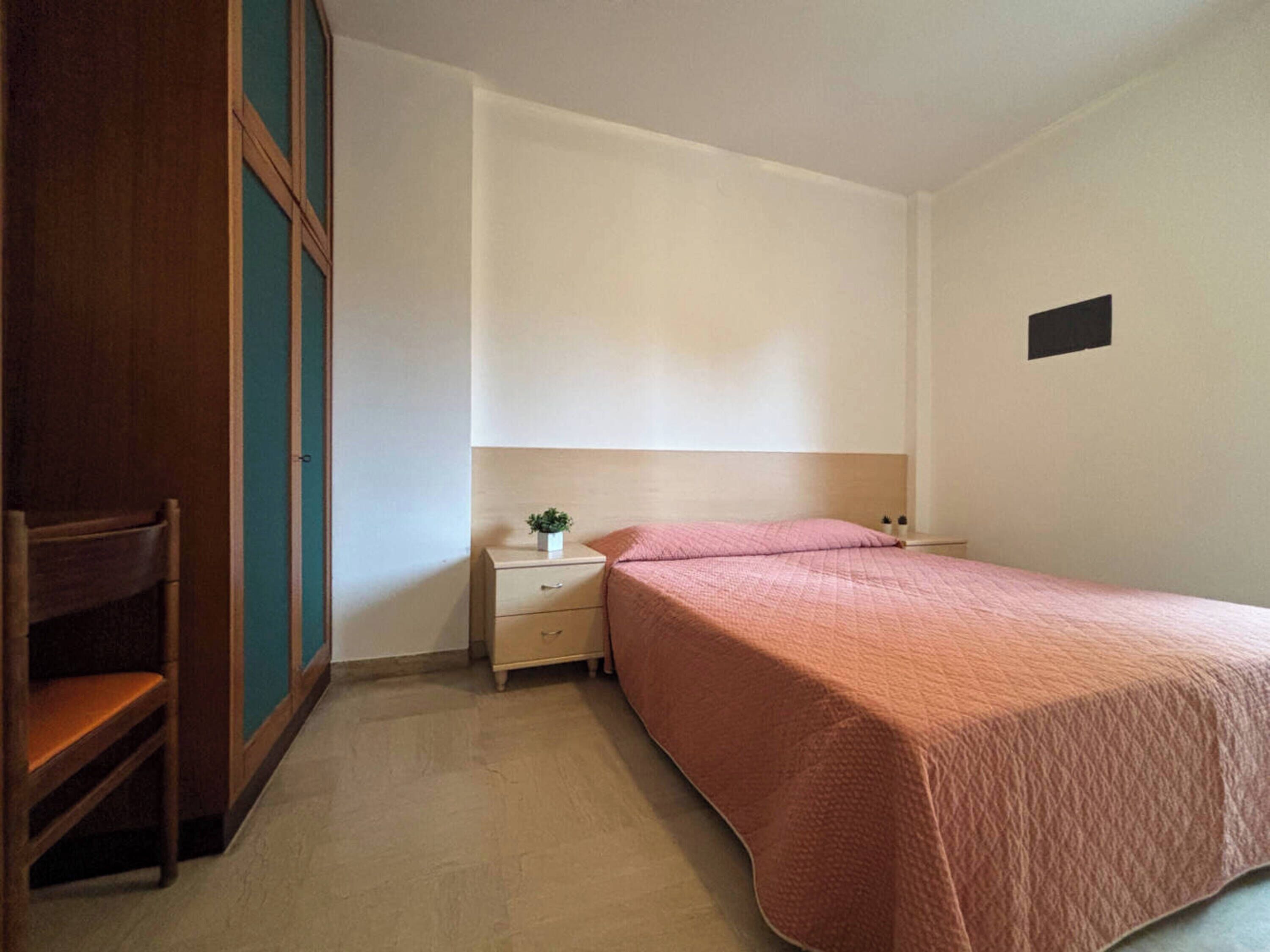 A bedroom with a bed, a nightstand, a small table with a plant, a chair, and a wardrobe.