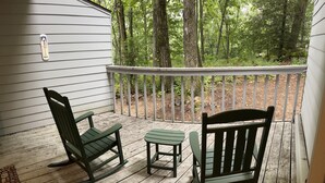 A wooden deck with two rocking chairs and a small table, surrounded by a white railing and a view of a forested area. - Ski-In/Ski-Out Comfy Condo (Wintergreen)