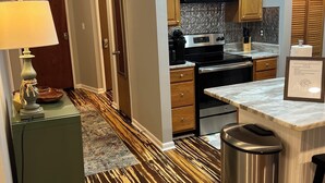 A modern kitchen with a microwave, stove, and wooden cabinets. A trash can and a bar stool are visible. - Ski-In/Ski-Out Comfy Condo (Wintergreen)