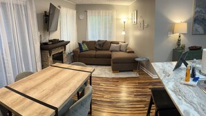 A modern living room with a wooden dining table, a brown sofa, a TV mounted on the wall, and a fireplace. - Ski-In/Ski-Out Comfy Condo (Wintergreen)