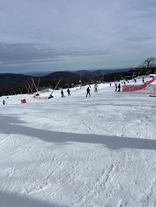 Una pista de esquí con esquiadores y snowboarders, senderos señalizados y una cadena montañosa distante.