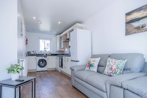 A modern kitchen with white cabinets, a washing machine, and a dining area with a grey sofa and a painting of a beach scene. - Swn Y Mor - 4 Bedroom - Llanelli (Llanelli)