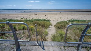 Vue sur la plage depuis une terrasse en bois dotée de rampes en métal, avec une plage de sable et des zones herbeuses.