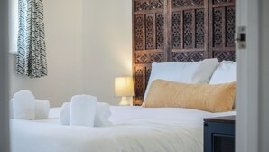 Un lit bien fait, avec du linge de lit blanc et un oreiller beige, une tête de lit en bois ornée de motifs complexes et un rideau à motifs à la fenêtre.