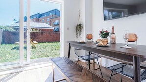 Un espace repas moderne avec une porte-fenêtre coulissante donnant sur un jardin, un téléviseur à écran plat fixé au mur, et une table dressée avec de la nourriture et des boissons.