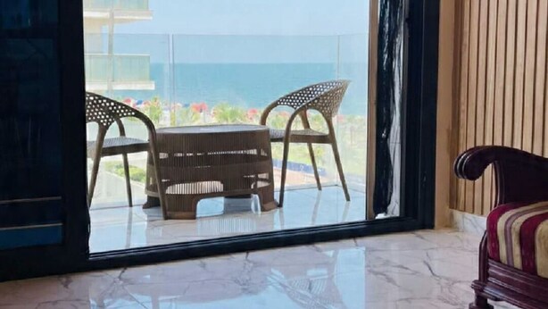 A room with a large sliding glass door leading to a balcony with two wicker chairs and a table. The floor is tiled with a glossy finish. - A Modern Haven Steps Away From the Beach (Al-Manasrah)