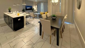 Cocina moderna con una isla central, comedor con mesa oscura y sillas blancas, y sala de estar con televisor y asientos.