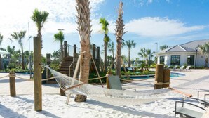 Zona de playa con una hamaca, palmeras, piscina y un edificio al fondo.