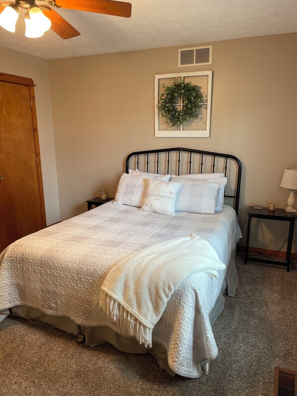 Une chambre à coucher comprenant un lit, une table de chevet, une lampe, un ventilateur de plafond, une porte et un tableau accroché au mur.