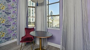 A room with a purple wall, a patterned wallpaper, a round table, a red chair, and a view of buildings through a window. - Hotel Fiona (San Francisco)