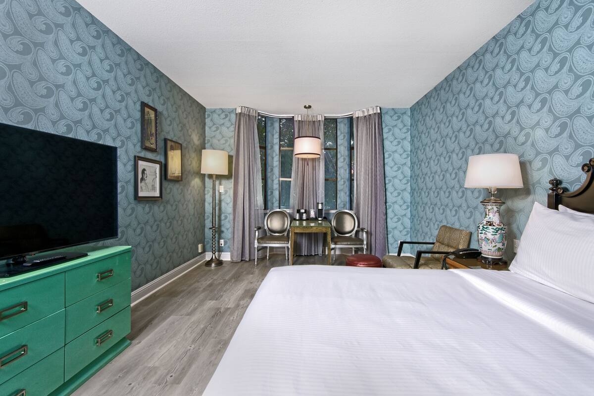 Habitación de hotel con una cama grande, un aparador verde, un escritorio con dos sillas, un televisor y una lámpara en un florero.