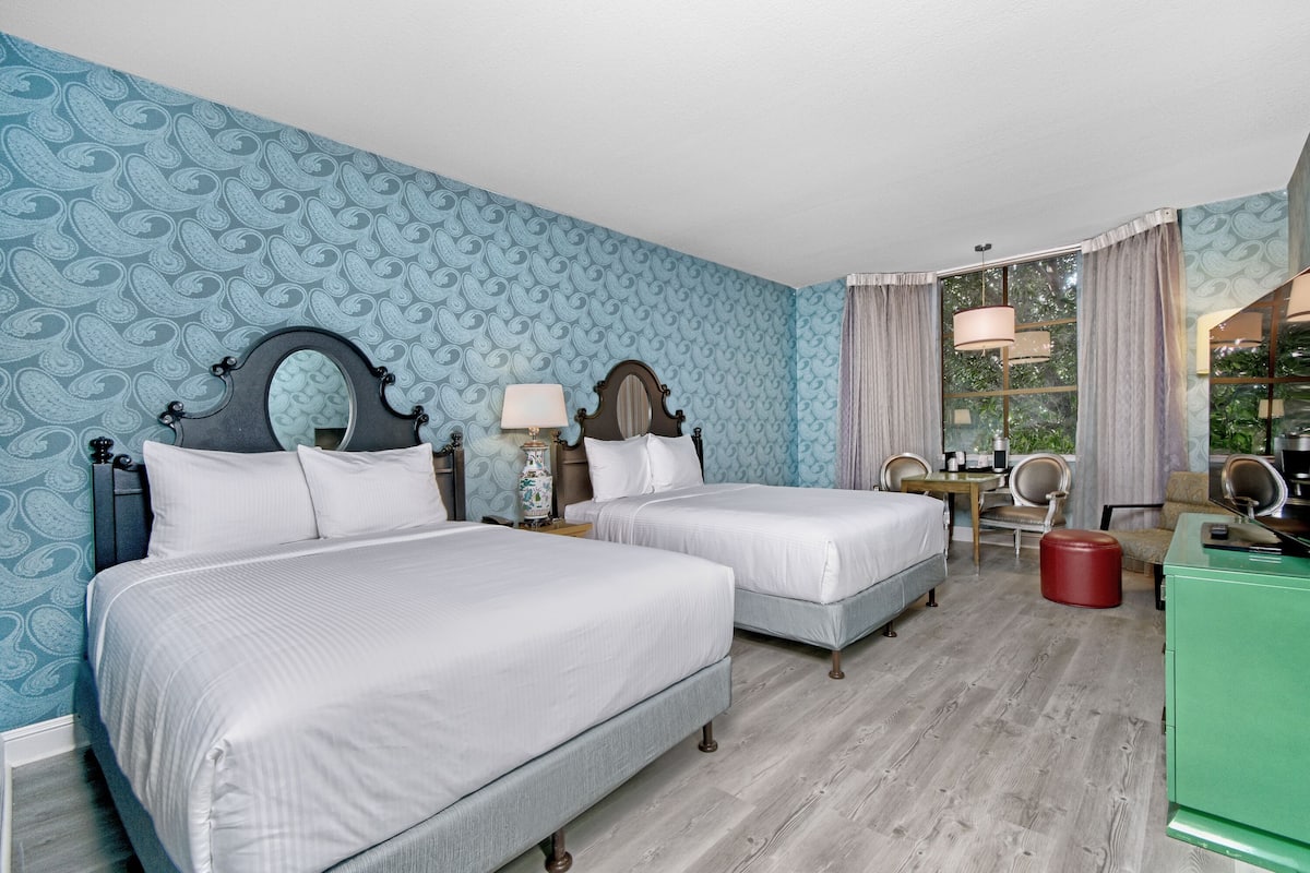 A hotel room with two beds, a desk, a chair, a mirror, and a window with curtains.