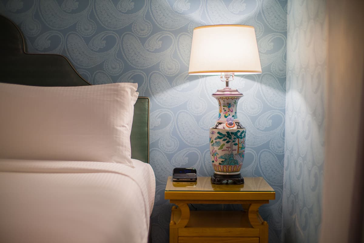 A neatly made bed with a pillow, a small bedside table with a lamp, and a mobile phone on it.