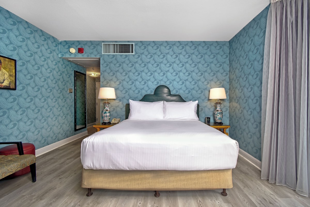 A hotel room with a large bed, two bedside tables with lamps, a red chair, and a door leading to another room.