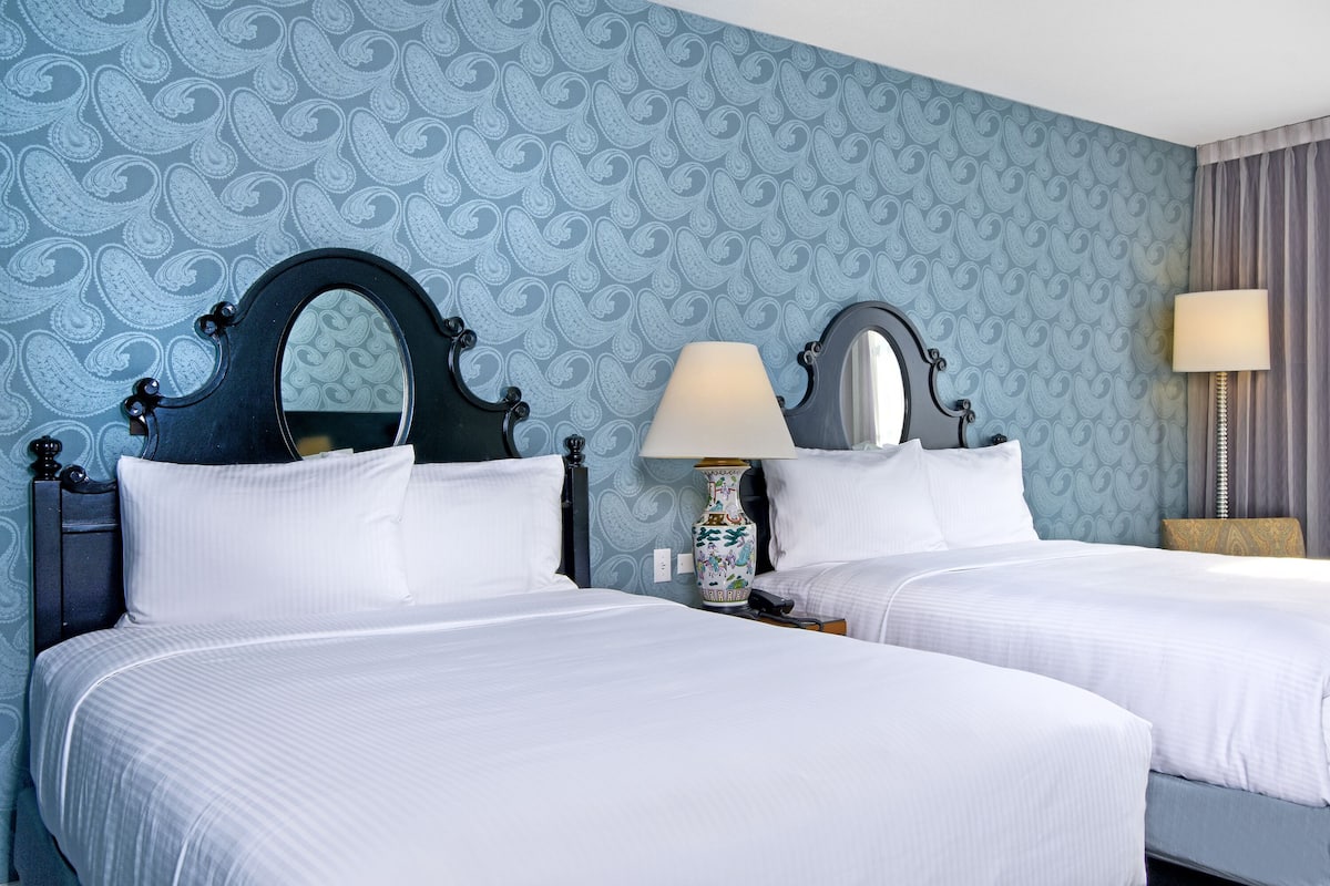 A hotel room with two beds, a large mirror above each bed, a vase on the nightstand, and a lamp on the side table.