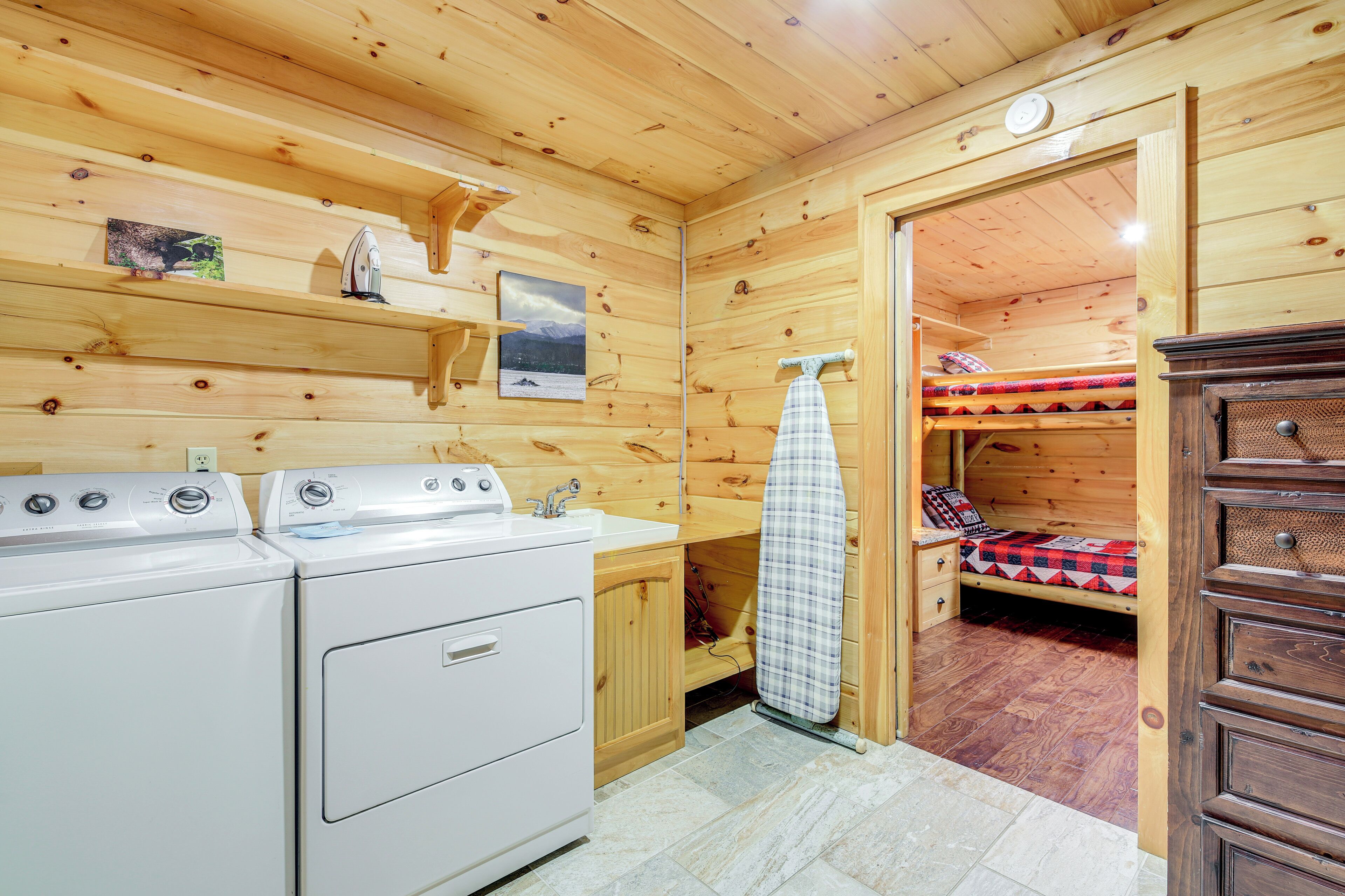 A compact wooden cabin with a washing machine, a bed with a red and black checkered blanket, and a small wooden cabinet.