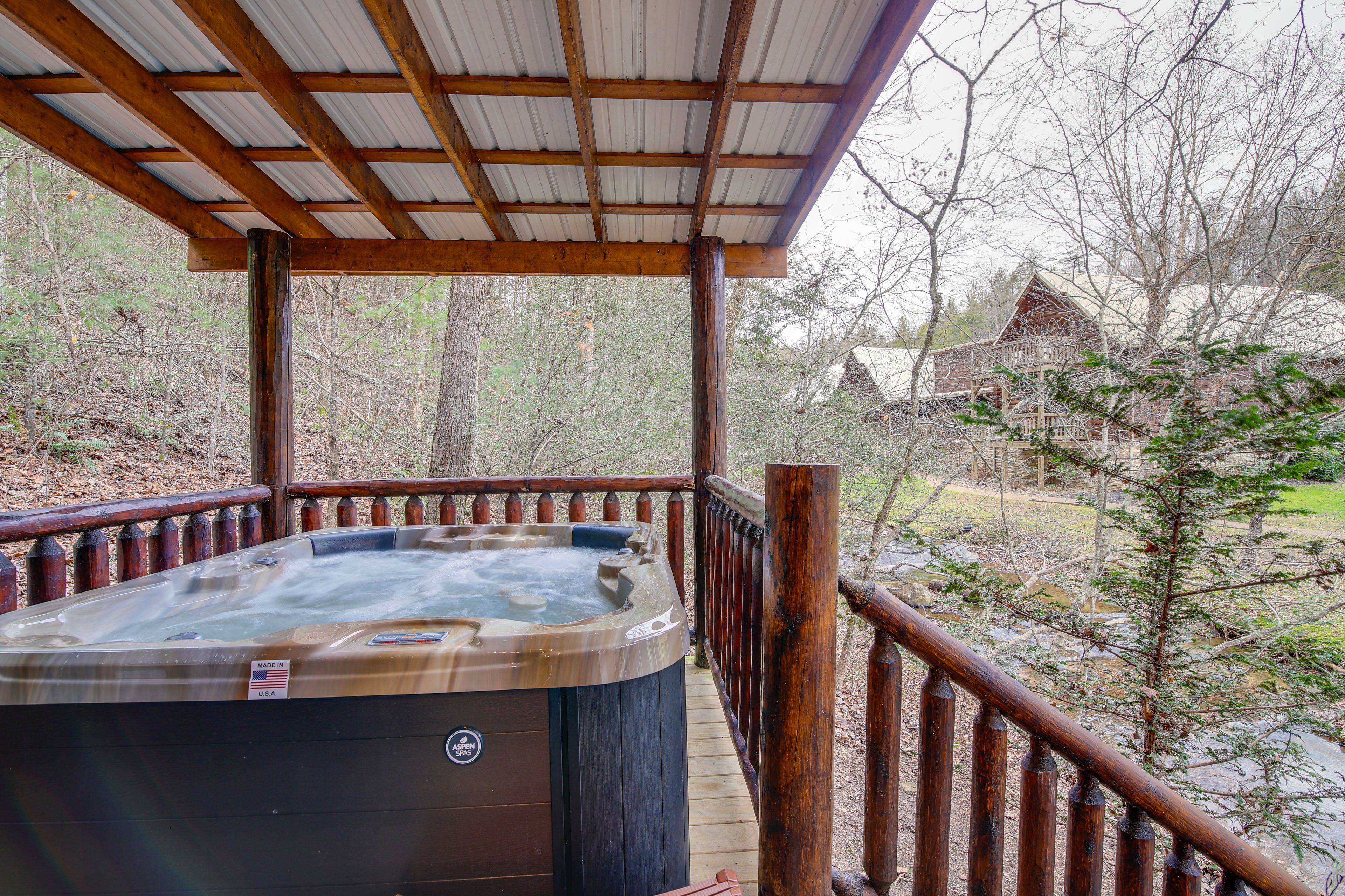 Un bain à remous sur une terrasse en bois, avec vue sur une forêt et une maison en arrière-plan.