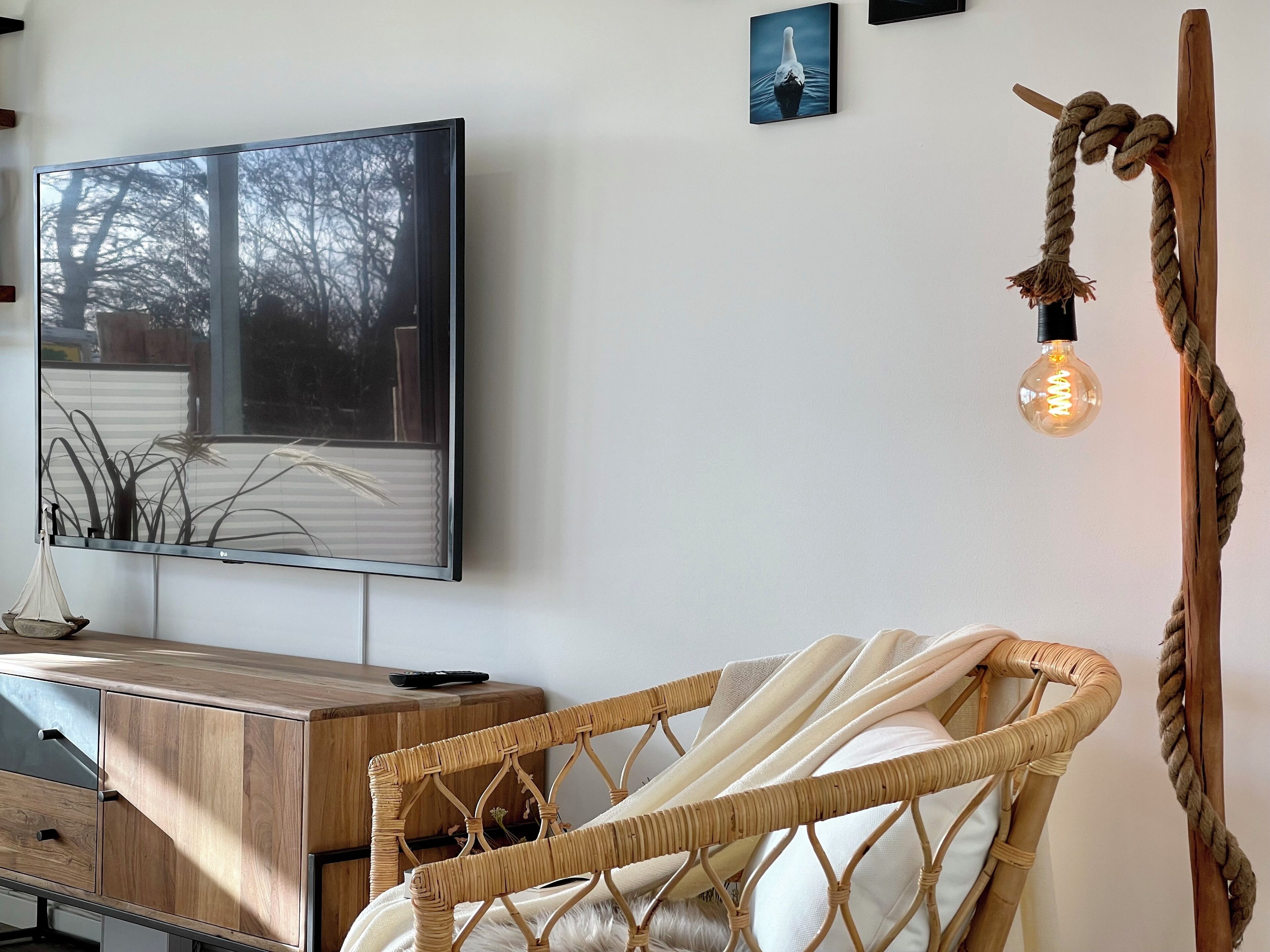 A room with a television mounted on the wall, a wooden cabinet with drawers, a wicker chair with a white cushion, and a rope decoration with a light bulb.