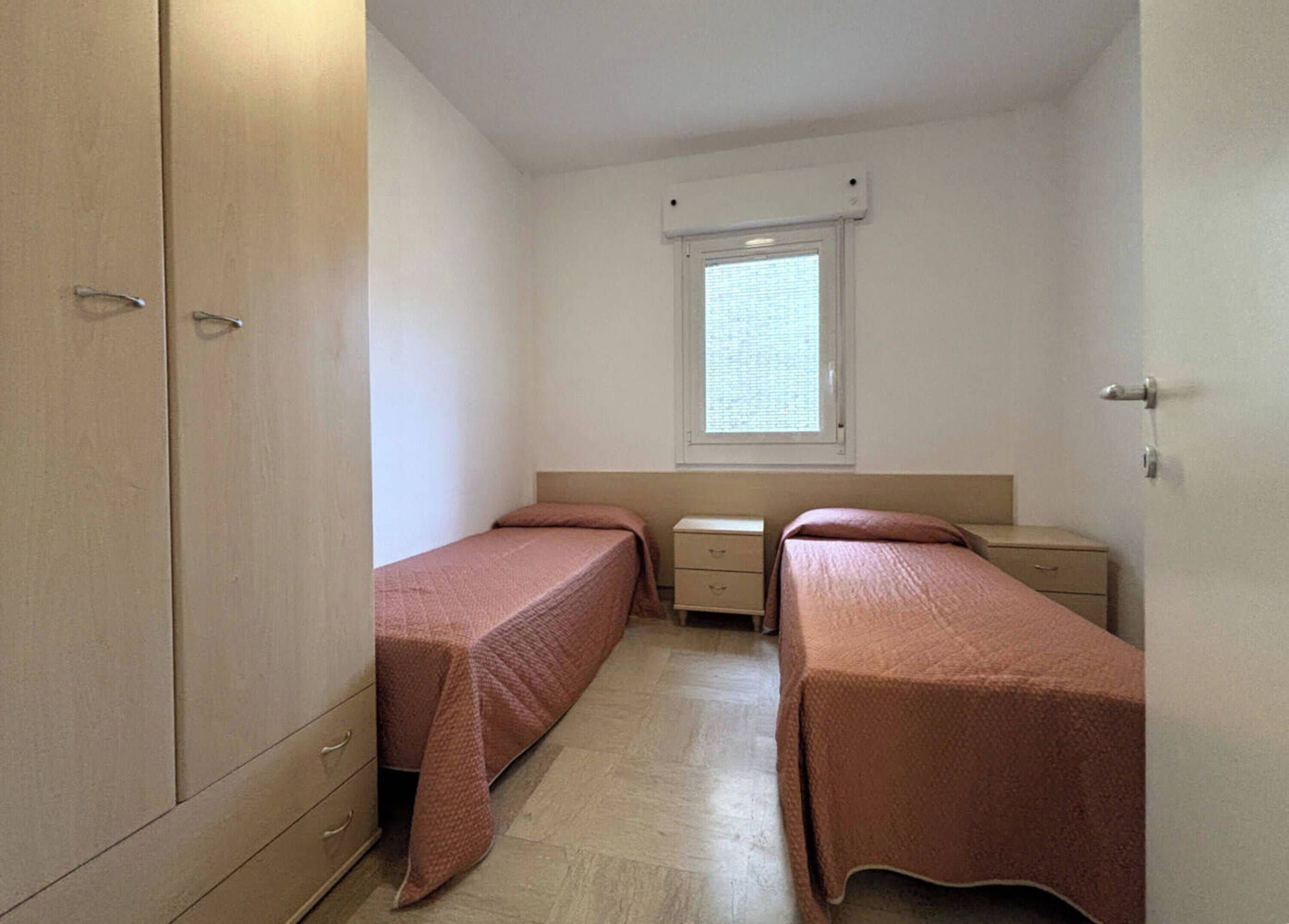 Una piccola stanza singola con due letti, una finestra, un armadio e un comodino.