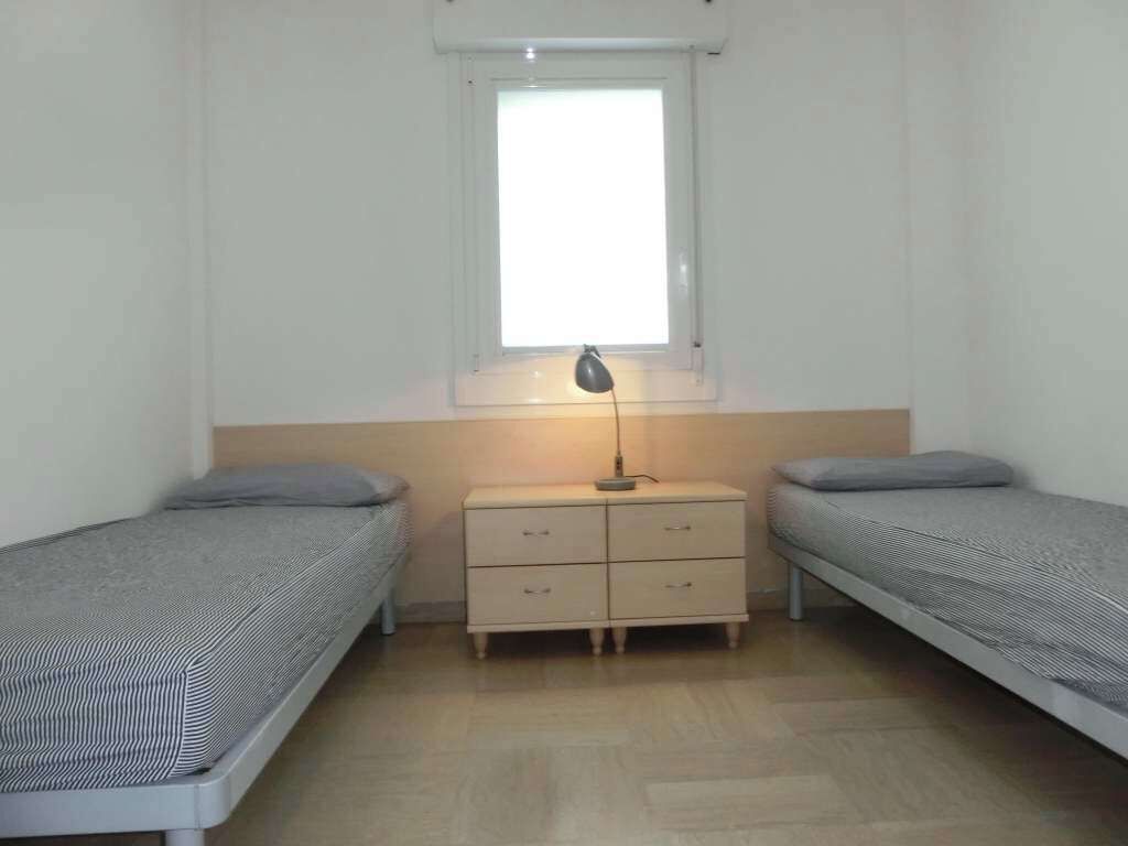 Un petit logement composé d’une seule pièce, comprenant deux lits simples, une table de chevet, une lampe et une fenêtre.