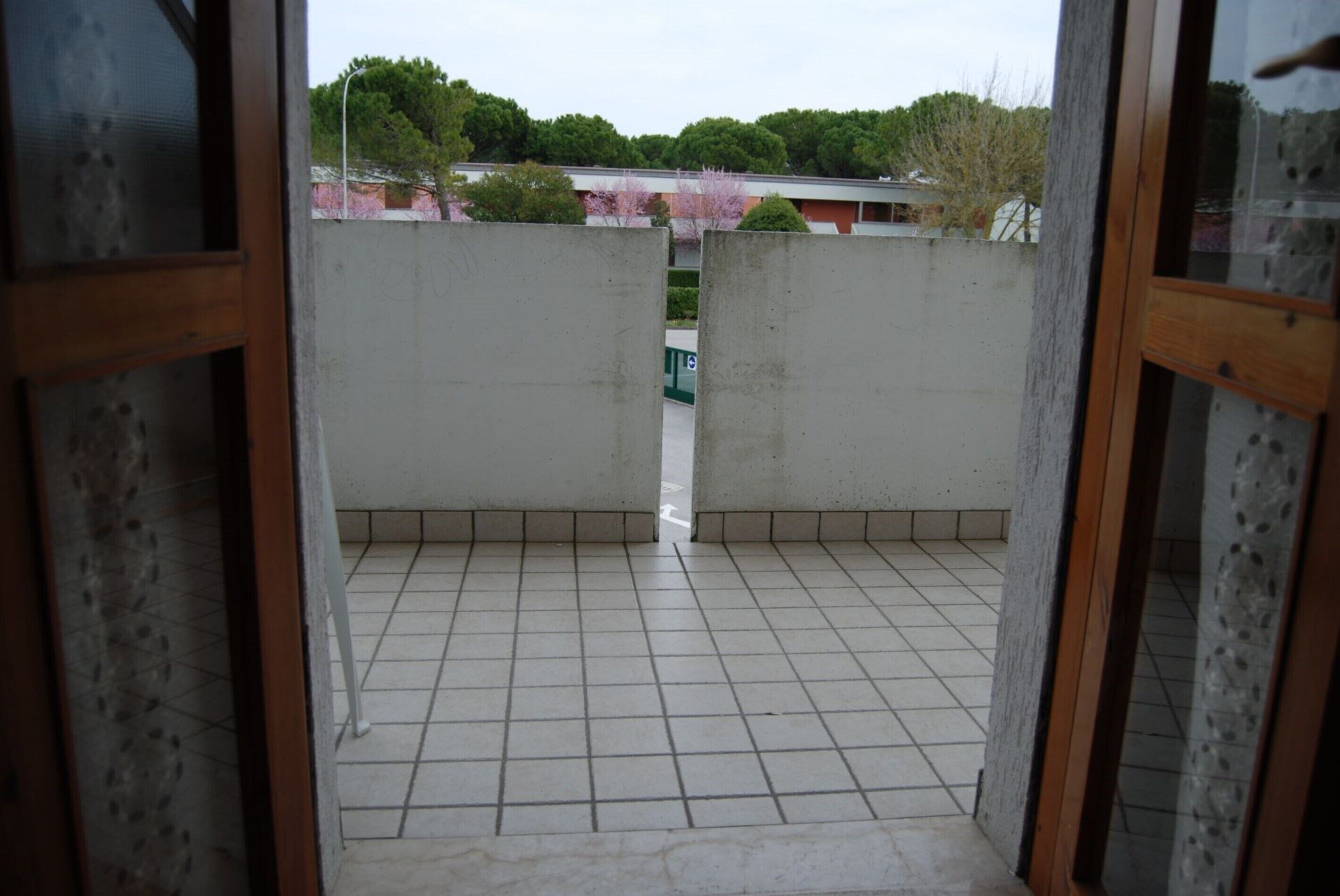 Une vue à travers une porte-fenêtre donnant sur un balcon, avec un sol carrelé, qui mène à une cour intérieure bordée d’une clôture verte et d’arbres.