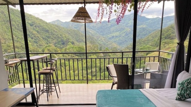 Een balkon met uitzicht op groene bergen, een zithoek met een tafel en stoelen, en twee bedden met blauwgroene beddengoed.