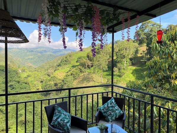 A balcony with a view of green hills, two wicker chairs with cushions, and hanging purple flowers.