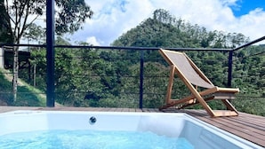 Een hottub met een houten terras en een lounge stoel, omgeven door weelderige begroeiing en een heldere lucht.