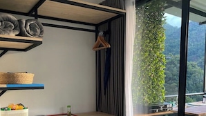 Een moderne kamer met een inbouw bureau, een kleine koelkast en uitzicht op een balkon met een houten terras en begroeiing.