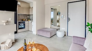 Un salon moderne avec un canapé, une table basse sur laquelle se trouvent des verres à vin et du fromage, un fauteuil en osier, et une cuisine en arrière-plan.