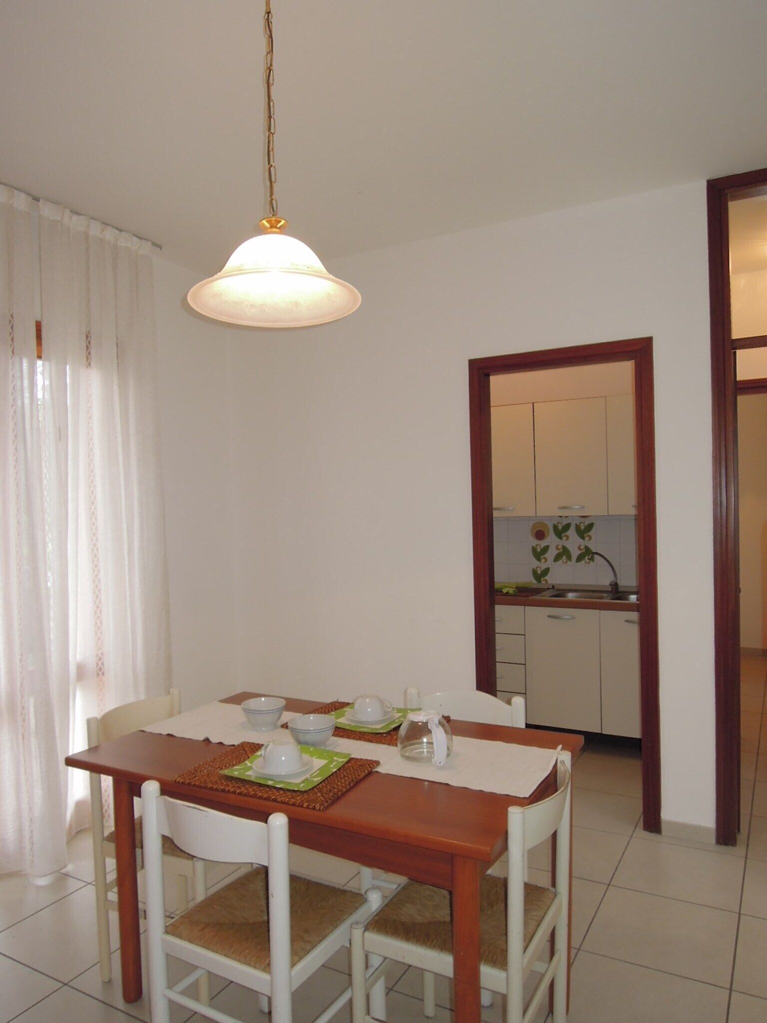 A dining area with a wooden table and chairs, a hanging light fixture, and a view into a kitchen with cabinets and a sink.