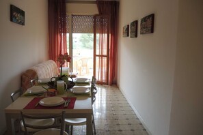 Un comedor con una mesa preparada para cuatro personas, sillas, un sofá y vistas a través de una ventana con cortinas.