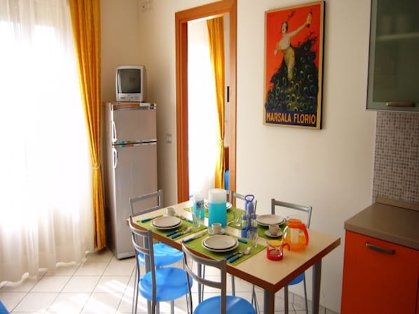 A dining area with a table set for four, a refrigerator, a television on top, and a poster on the wall.