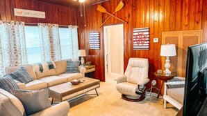 Ein gemütliches Wohnzimmer mit Holzbalkendecke, einem beigefarbenen Sofa, einem weißen Sessel, einem Fernseher und einem kleinen Tisch mit einer Lampe.