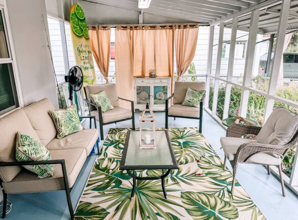 A sunlit patio with a sofa, chairs, and a table, featuring a tropical leaf-patterned rug and a decorative lantern.