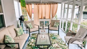 Eine sonnendurchflutete Terrasse mit einer Couch, Stühlen und einem Tisch, mit einem Teppich mit tropischen Blätermotiven und einer dekorativen Laterne.