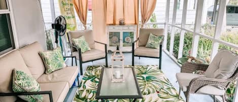 A sunlit patio with a sofa, chairs, and a table, featuring a tropical leaf-patterned rug and a decorative lantern.
