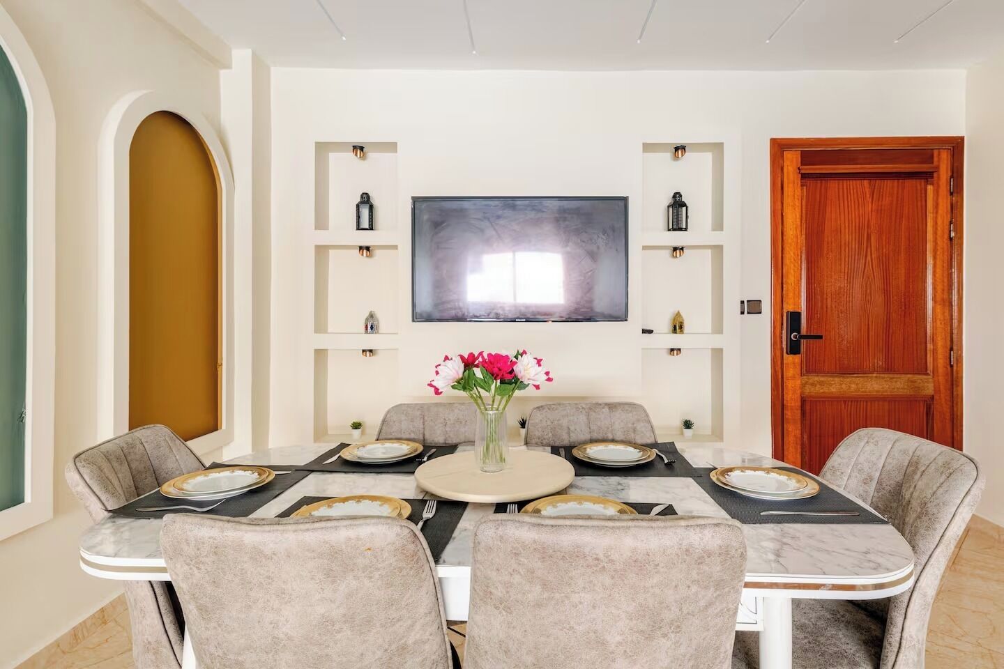 A dining room with a large table, six chairs, a vase with flowers, a television on the wall, and a wooden door.