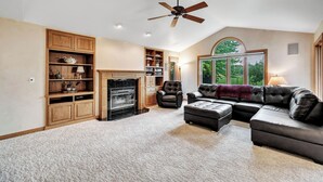 A spacious living room with a fireplace, leather furniture, wooden shelving, and a large window offering a view of greenery.