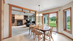 A spacious dining area with a wooden table and chairs, a fireplace, and a view of a green landscape through large sliding glass doors.
