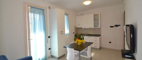 A modern living room with a blue sofa, a dining table with chairs, a kitchen area with white cabinets, and a TV mounted on the wall.
