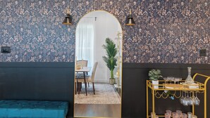 Una habitación con papel tapiz floral, un espejo, un sofá azul, una zona de comedor con mesa y sillas, y una barra con vasos y botellas.