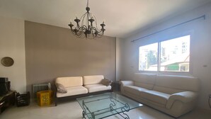 Una sala de estar con dos sofás blancos, una mesa de centro de vidrio, una araña y un ventanal con vista a la ciudad.