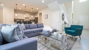 A modern living room with a grey sofa, a teal armchair, and a glass coffee table. - Amazing Home at Windsor Westside (Kissimmee)