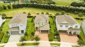 Área residencial com três casas de dois andares, cada uma com garagem, cercada por gramados bem cuidados e uma fileira de palmeiras.
