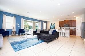 Uma sala de estar espaçosa com um sofá seccional cinza escuro, um tapete azul e uma cozinha com armários de madeira e uma geladeira de aço inoxidável.
