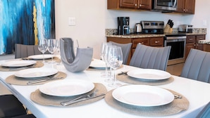 A dining table set with plates, cutlery, and wine glasses, with a modern kitchen in the background. - Charming Home With Private Pool at Encore Resort (Kissimmee)