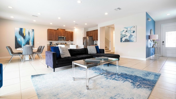 A modern living room with a glass coffee table, a sofa with cushions, a dining area with chairs, and a kitchen in the background. - Charming Home With Private Pool at Encore Resort (Kissimmee)